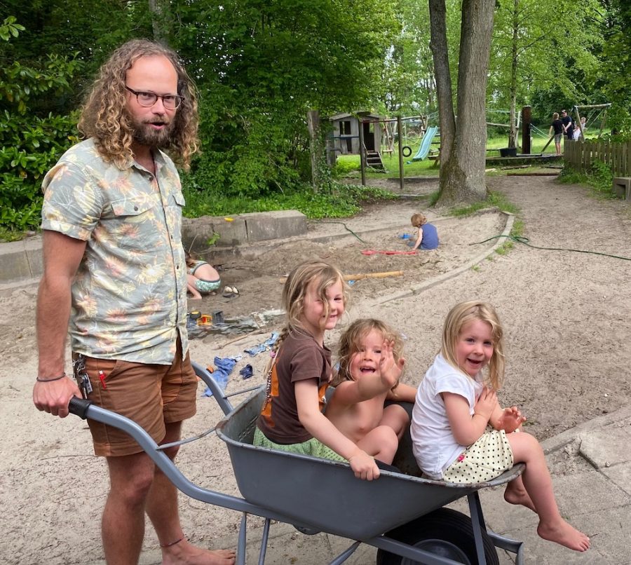 Portret van Tobias Hoendervangers, staflid op democratische school De Ruimte in Soest. Hij heeft een kruiwagen vast met daarin drie peuters die lachen.