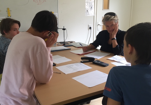 Op de rug gezin zie je drie leerlingen van democratische school De Ruimte aan een tafel, met werkbladen voor zich. Rijk, docent op De Ruimte, kijkt hen geïnteresseerd aan