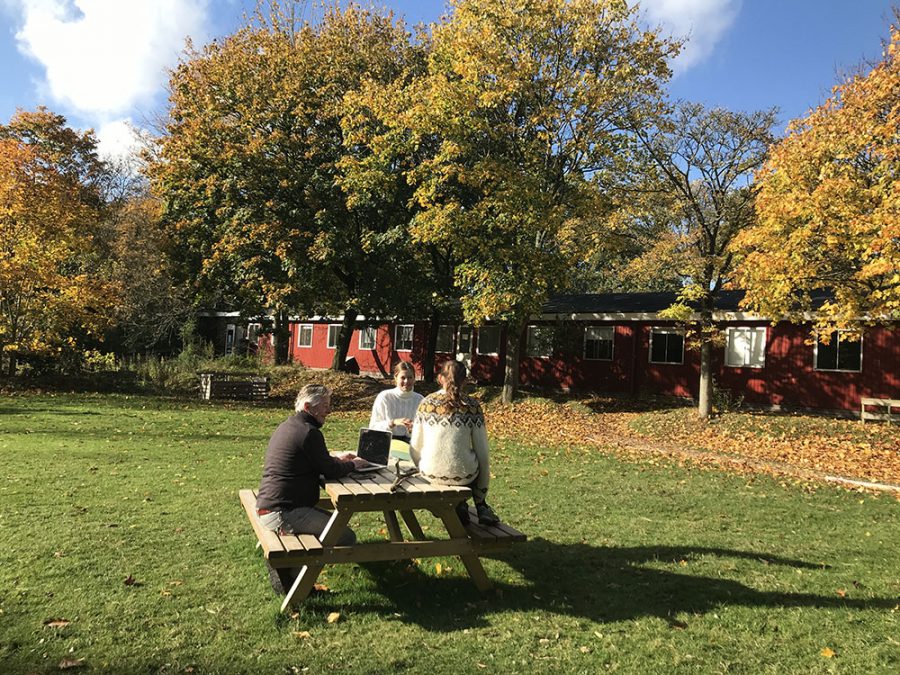 A staffmember and two students are sitting at a picknick table, outside democratic school De Ruimte. Behind them there are autumn trees and a low, red building