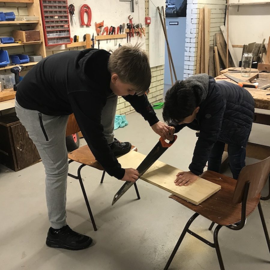 One student is trying to saw a plate of wood. Another student is helping bu holding the saw with his two hands