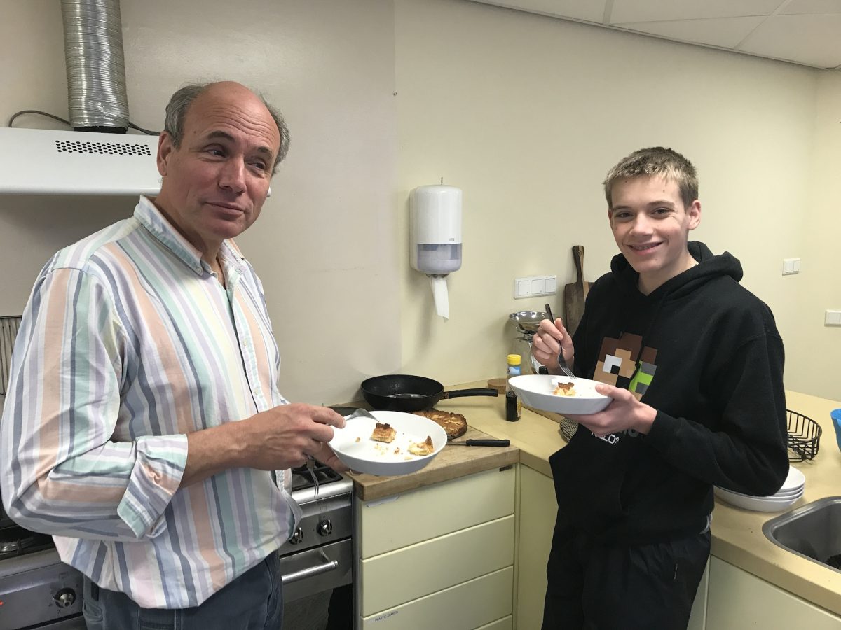 Docent Frithjof en Robin in de keuken op democratische school De Ruimte. Links staat Frithjof, hij heeft een schoteltje met taart vast en met zijn andere hand wil hij een stukje taart prikken. Robin staat rechts en heeft ook een schoteltje met taart in zijn handen. Hij kijkt blij.