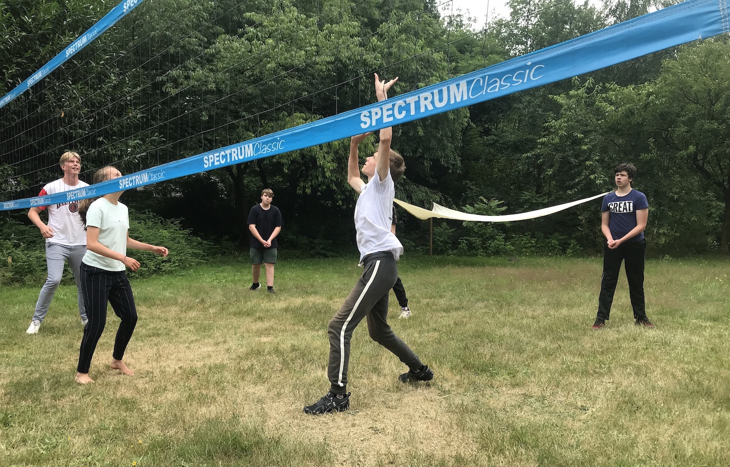 Six people stand behind a blue volleyball net, on a lawn. In the middle is Robin, student at De Ruimte, he looks up to play a ball back