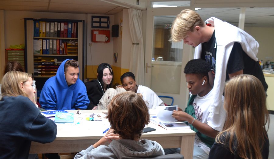 Seven pupils at De Ruimte are sitting around a table in domain 3. They are watching one of them looking in a book on the table. Another student is standing behind him and also looks in the book.