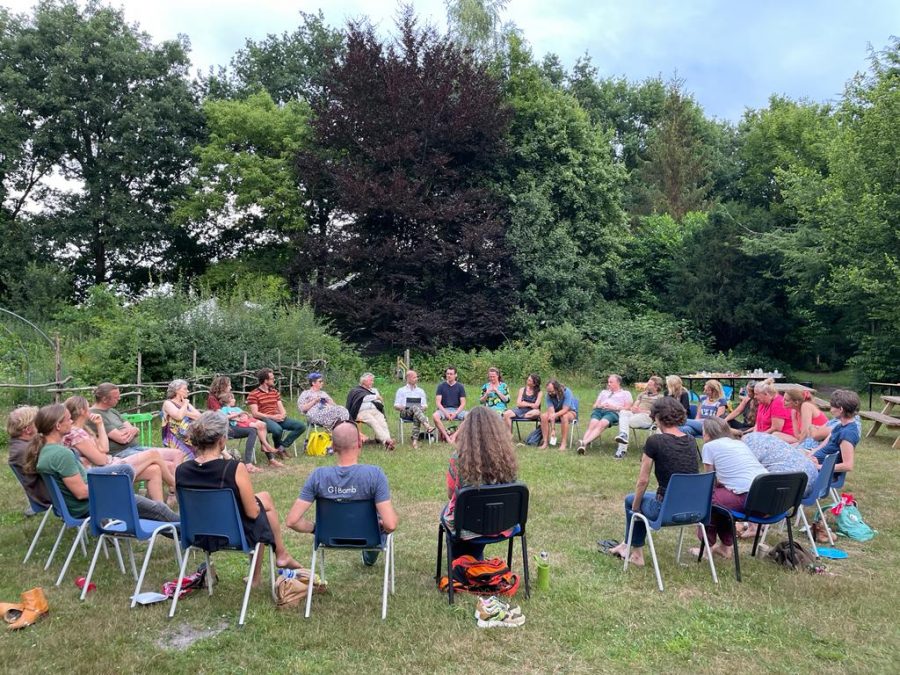 Ouders van democratische school De Ruimte zitten buiten in een kring voor de ouderkring. Ze zitten op stoelen in het gras, achter hen zijn bomen te zien.