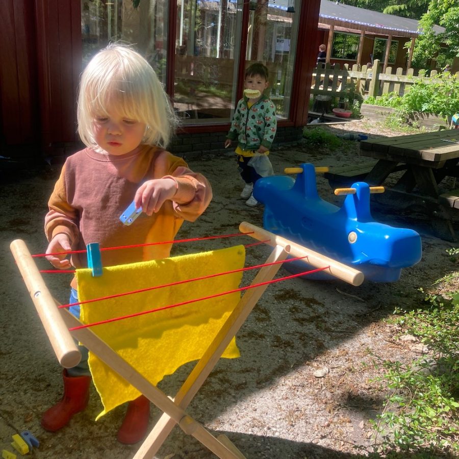 Een peuter staat voor een houten speelwasrekje. Er hangt een geel doekje aan dat ze met een knijper vast wil zetten