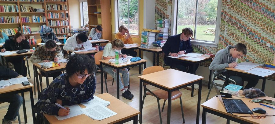 Een stuk of tien studenten zitten verspreid aan tafels in een lokaal. Ze zitten gebogen over examens en zijn aan het schrijven.