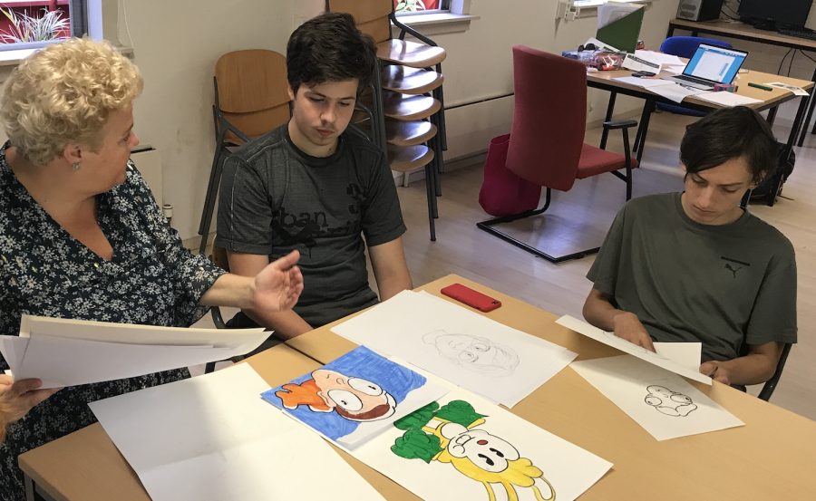 The teacher at the left gestures to the pupil next to her. Colorful drawings are on the table. Another student watches a paper in his hands.