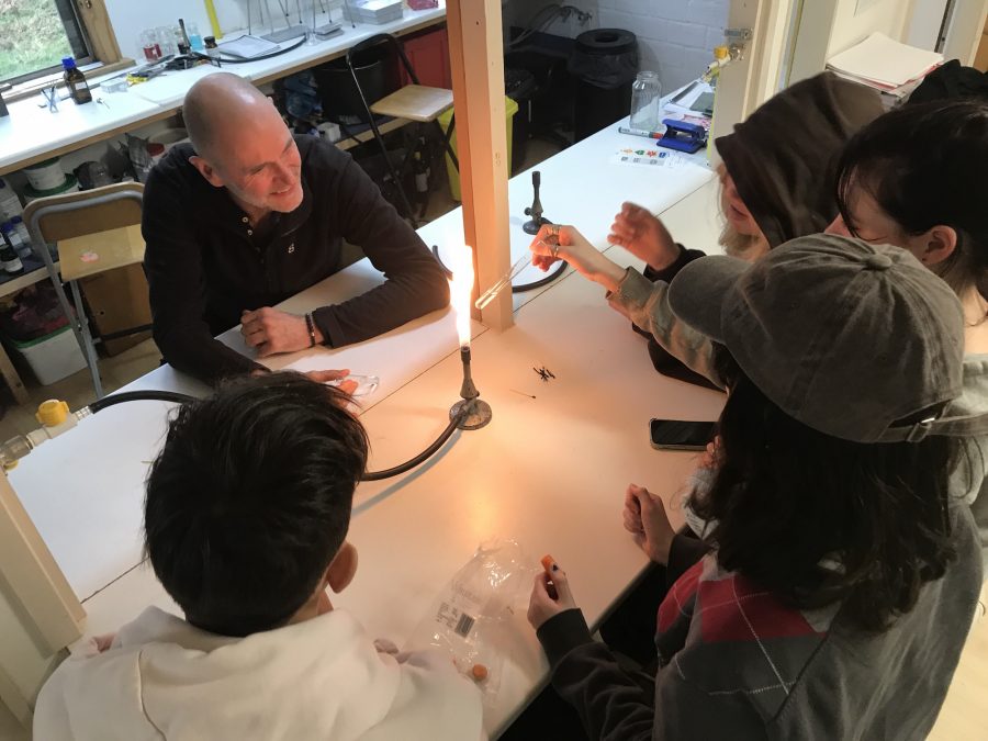 Op de rug gezien zitten vier leerlingen aan een tafel in het scheikundelab, ze kijken naar een gasbrander met een vlam. Een van hen houdt een reageerbuis in de vlam. De docent kijkt lachend, zijn gezicht licht op door de vlam.