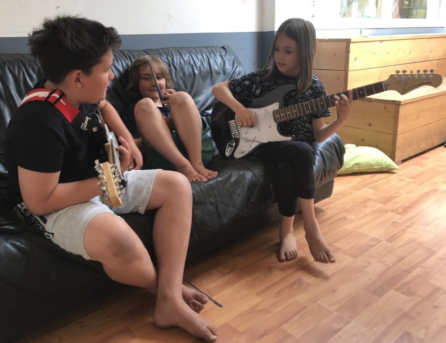 three young pupils are sitting on a couch in a hallway. The ones on the left and right both hold an electric guitar, the one in the middle holds a drumstick to his nose.