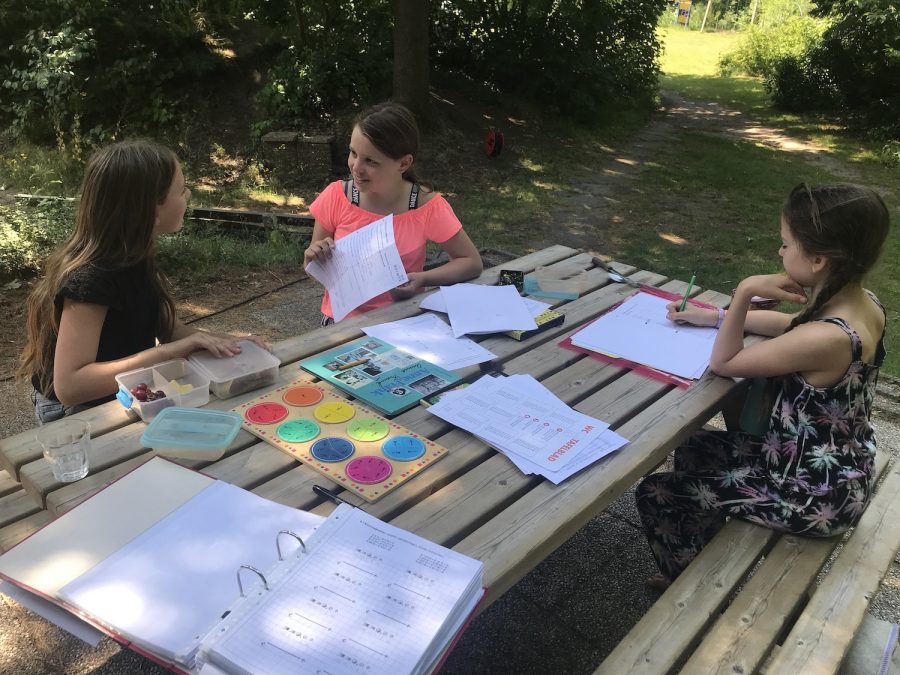 drie leerlingen van democratische school De Ruimte zitten buiten aan een picknicktafel. Op tafel liggen werkbladen, een map en een breukenplank