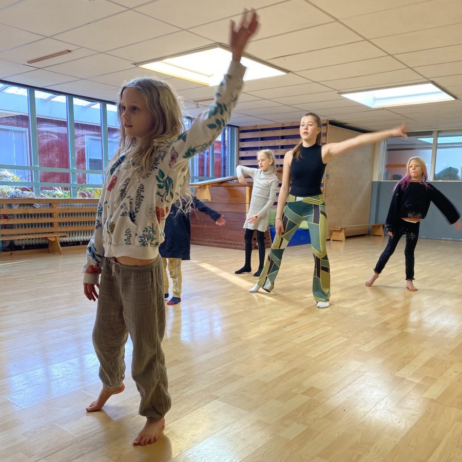 Five pupils are dancing, they look at themselves in the mirror (which is not on the picture). Four of them are younger than the fifth, who is guiding the dance.