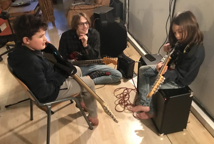 Two younger students left and right are sitting on a chair and an amplifier. They hold electric guitars in their hands. The older student in the middle sits on the floor and watches smiling