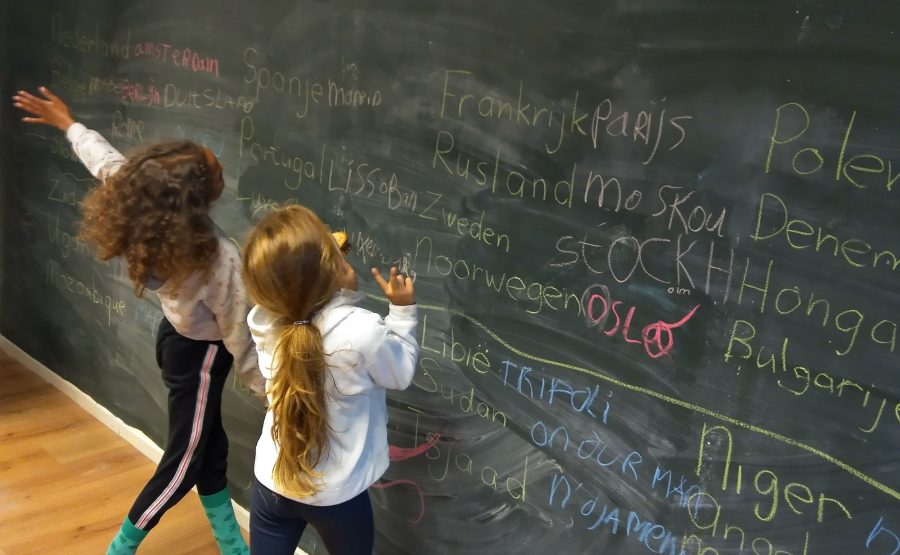 Twee jongere leerlingen staan voor een zwarte krijtwand, in domein 1. Op de muur staan namen van steden met krijt geschreven in verschillende kleuren.