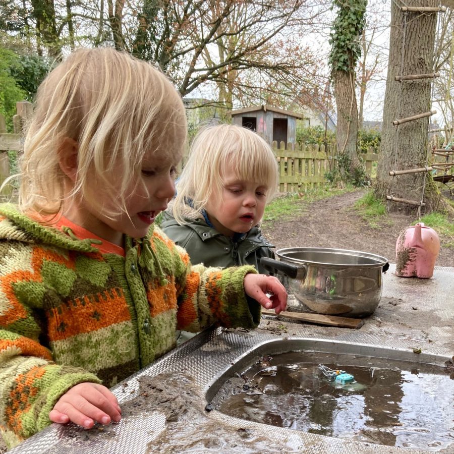 twee peuters staan buiten bij een aanrecht. Er ligt veel zand op het aanrechtblad.. De wasbak staat vol water. Ze kijken naar een autootje dat in het water ligt.