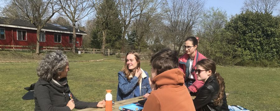 links op de foto zit een docent Engels aan een picknicktafel, rechts aan de tafel zitten vier studenten. Ze zitten buiten, achter hen is een deel van het rode schoolgebouw te zien, gras en bomen.