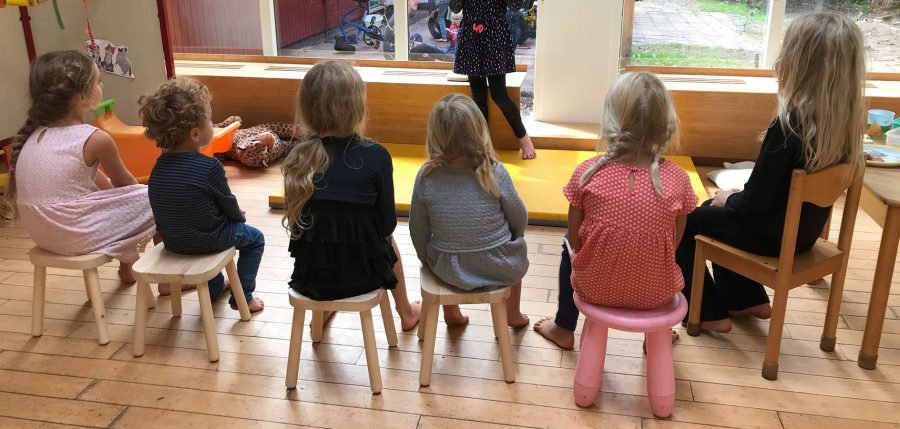 toddlers are sitting on small stools in a row. They're seen on the back. They are watching another student playing an act