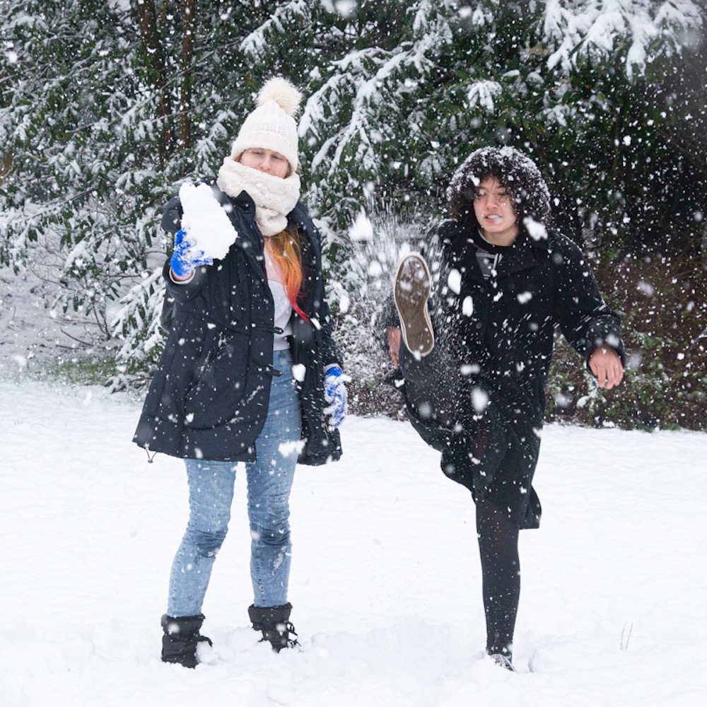 Twee studenten in de sneeuw, een van hen schopt tegen een sneeuwbal die uit elkaar spat