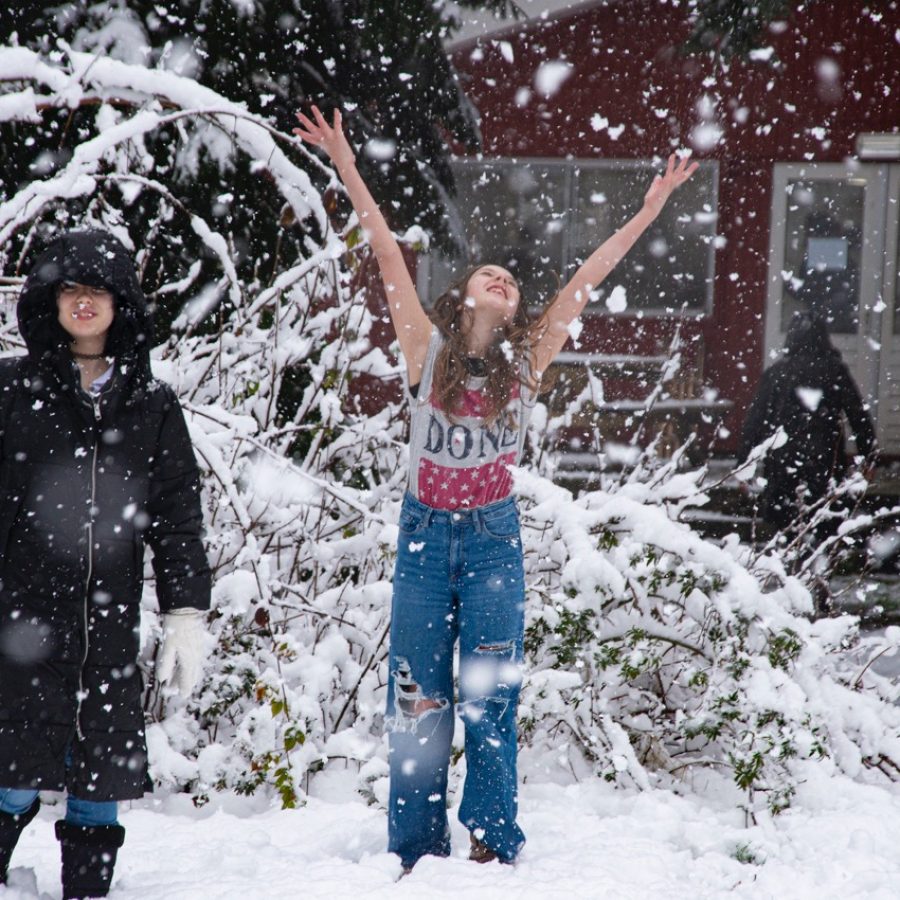 Twee studenten staan in de sneeuw, een van hen heeft alleen een T-shirt aan en reikt met haar armen naar boven, haar gezicht richting de vallende sneeuw, ze kijkt blij