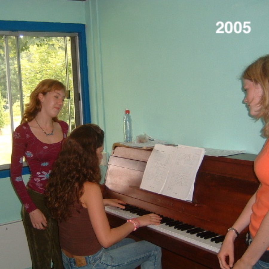 Een student achter de piano en twee studenten staan erbij, ze lachen.