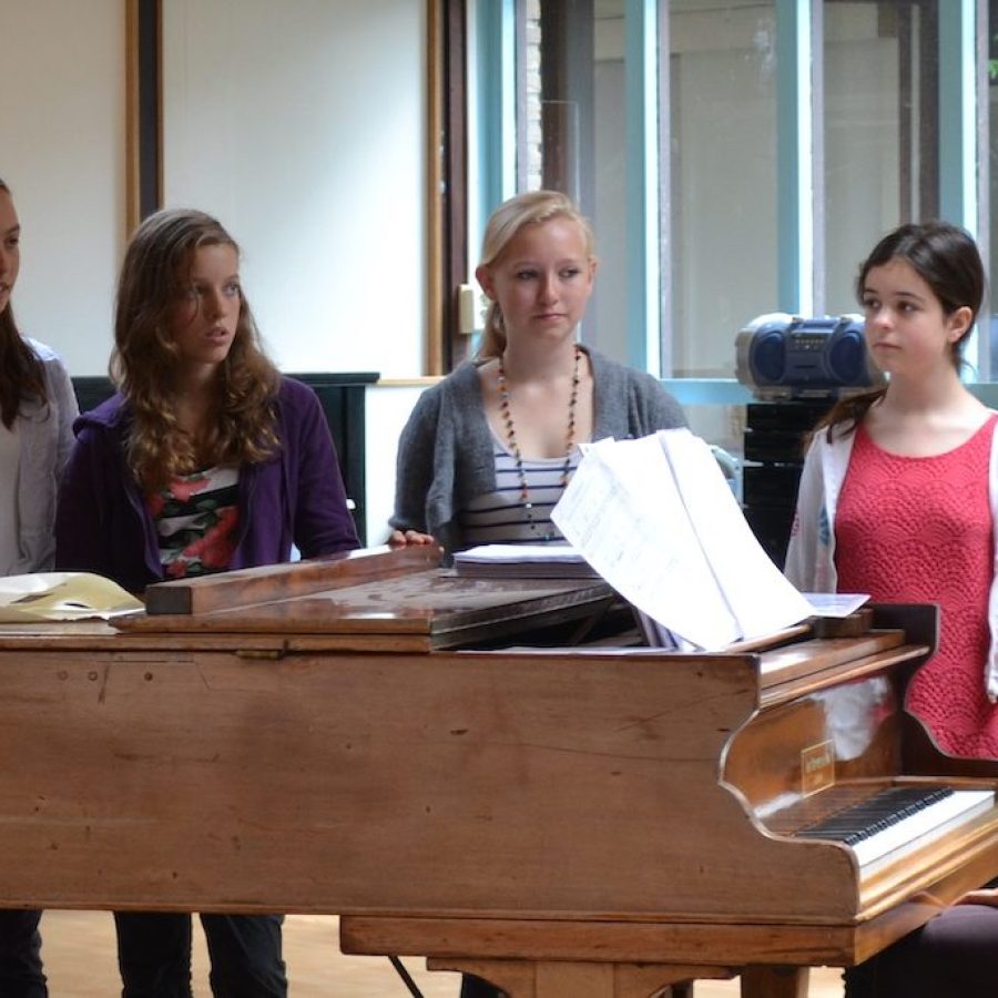 Zangdocent Meike van de Linden achter de piano, studenten kijken toe, klaar om te gaan zingen.