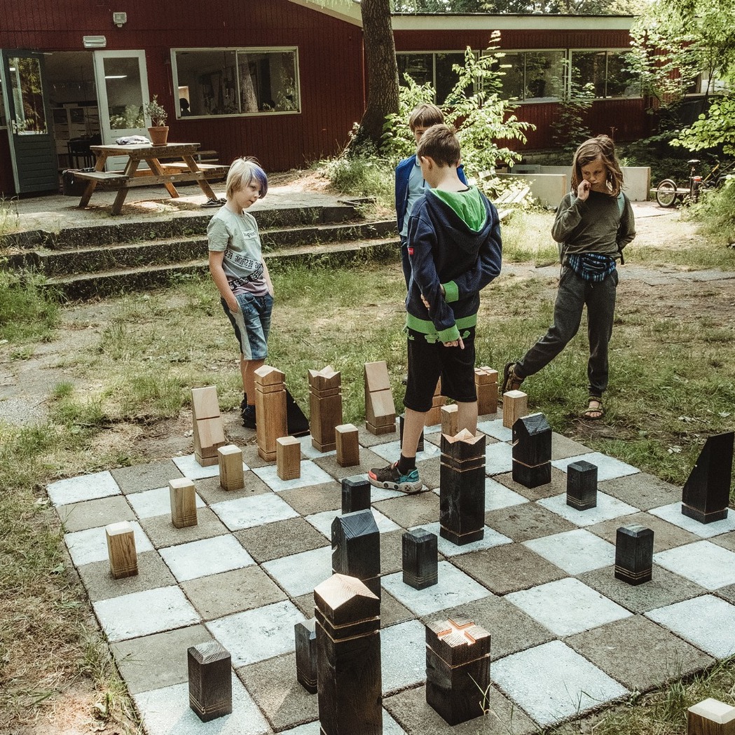 Vier studenten schaken buiten op een groot schaakbord, gemaakt van tegels en grote houten schaakstukken.