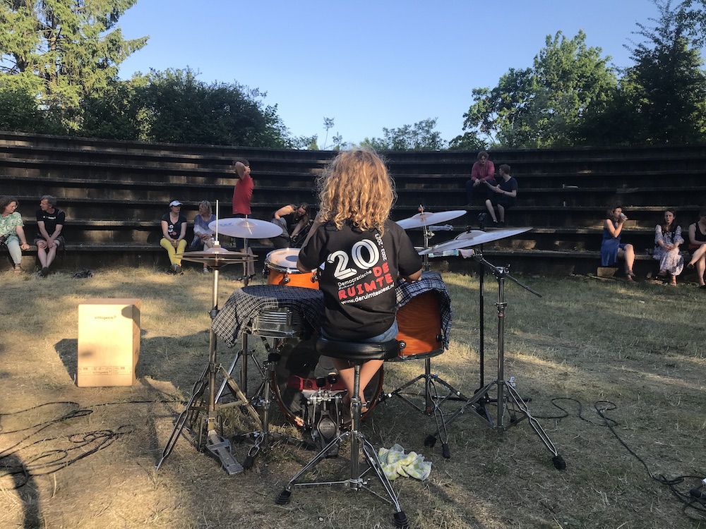 Een jong mens op de rug gezien, achter een drumstel. Hij draagt een T-shirt met het logo van 20 jaar De Ruimte. Op de achtergrond het amfitheater