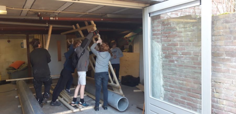 Studenten aan het werk in de kelder, ze tillen samen het houten geraamte omhoog van de zeilboot