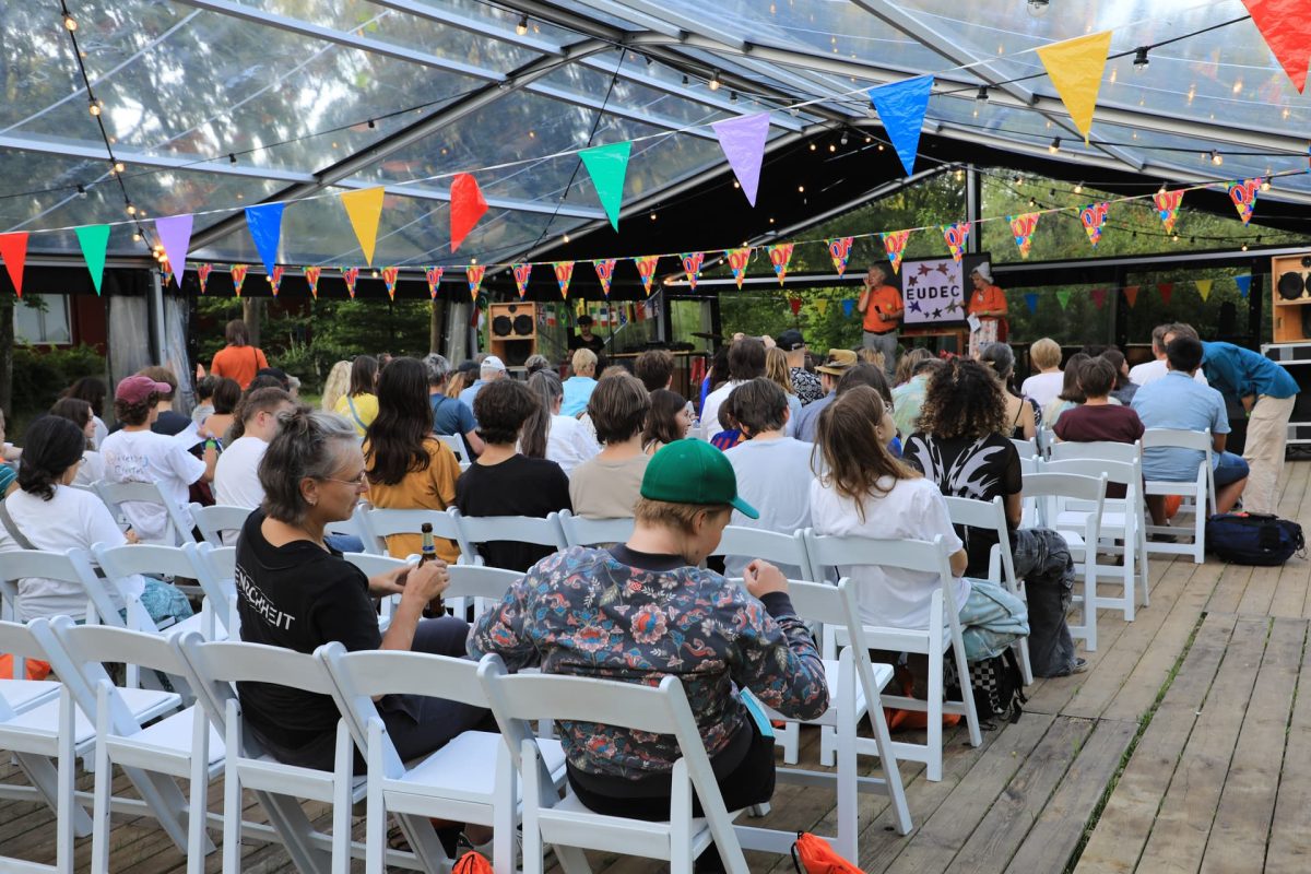 Rijen witte stoelen met publiek, in een doorzichtige tent versierd met vlaggetjes. Op het podium staan twee presentatoren in het Oranje.