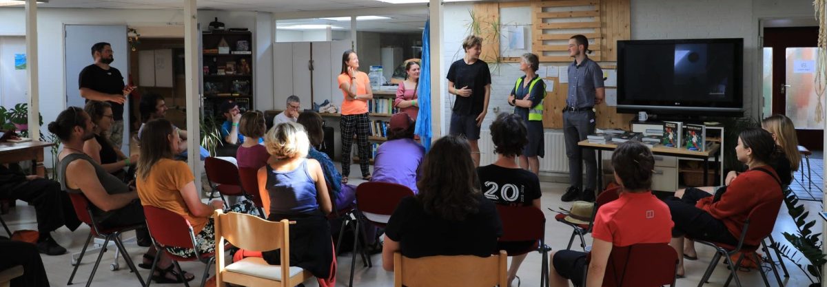 Publiek zit op stoelen in een zaal. Ze kijken naar vijf mensen die iets vertellen.