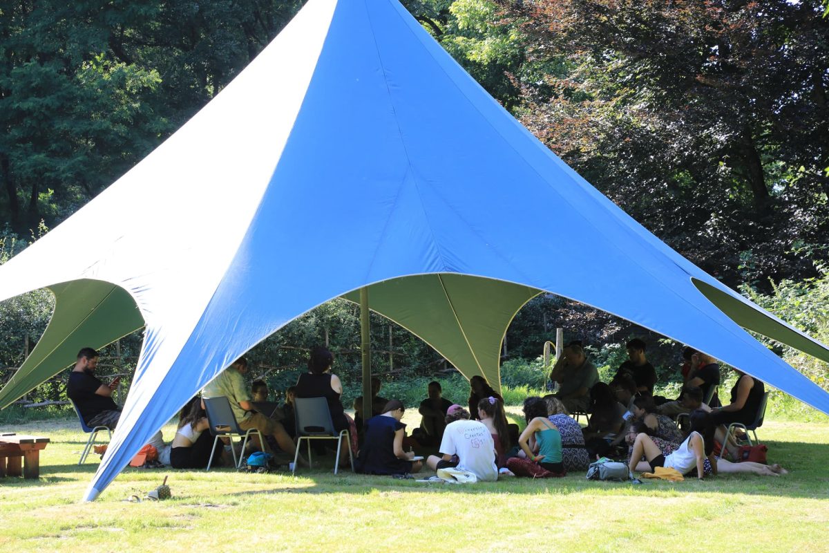 Een blauwe piramidevormige tent staat op het gras. Onder de tent zitten mensen in een kring.