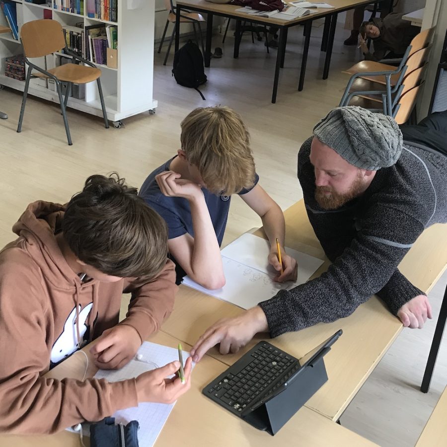 20250626_vacature-docenten Twee studenten en een staflid van democratische school De Ruimte zijn bezig met rekenen. De studenten zitten aan tafel, het staflid ligt op tafel en wijst met zijn vinger naar een van de schriften.