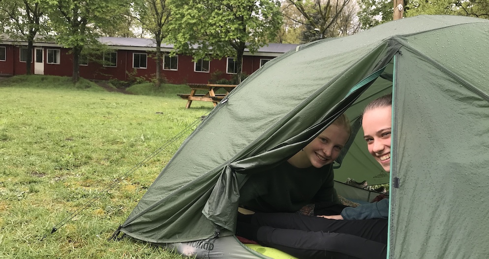 Twee studenten van democratische school De Ruimte kijken vanuit hun tent lachend naar de camera. De tent staat op een grasveld, een eindje daarachter staat het schoolgebouw.